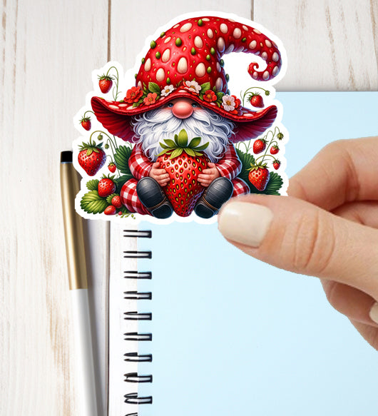 Sticker of a gnome with a strawberry hat held by a hand next to a notebook.