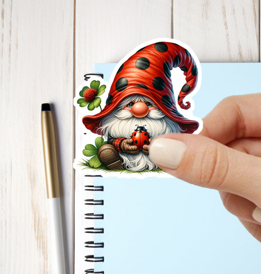 Hand holding a sticker of a gnome with a red hat and a ladybug, next to a pen and notebook on a light wooden surface.