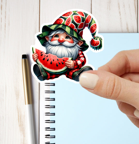 Sticker of a gnome with watermelon theme held by a hand next to a notebook and pen.