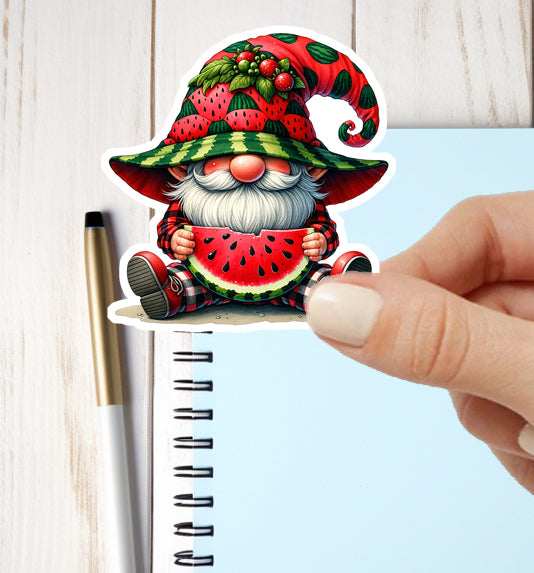 Gnome sticker with watermelon design held by a hand next to a notebook