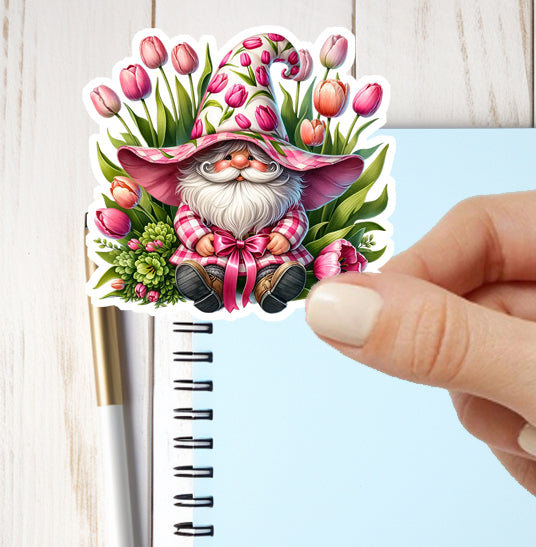 Sticker of a gnome with a pink hat and flowers held by a hand next to a notebook.