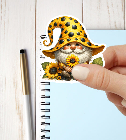 Hand holding a sticker of a gnome with a sunflower against a notebook background
