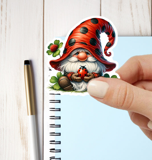 Hand holding a sticker of a gnome with a red hat and a ladybug, next to a notebook and pen.