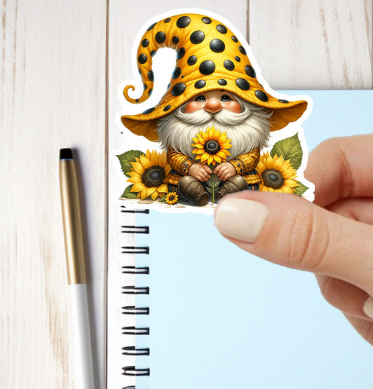 Hand holding a sticker of a gnome with sunflowers next to a notebook and pen.
