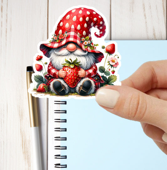 Hand holding a sticker of a gnome with a red hat and strawberry, placed on a notebook.