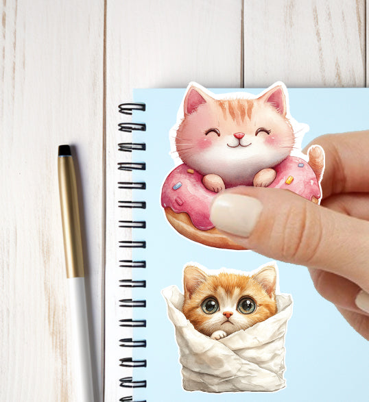 Hand holding a cute cat-shaped donut magnet next to a notebook with another cat magnet on a light wooden surface.