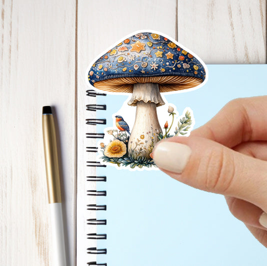 Hand holding a decorative mushroom sticker over a notebook with a pen on a wooden surface.