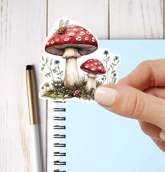 Sticker of red mushrooms with white spots held over a notebook with a pen on a light wooden surface.