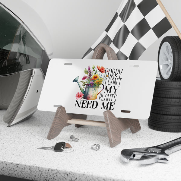 License plate with floral design and text 'Sorry I can't my plants need me' on a stand, with car parts in the background.