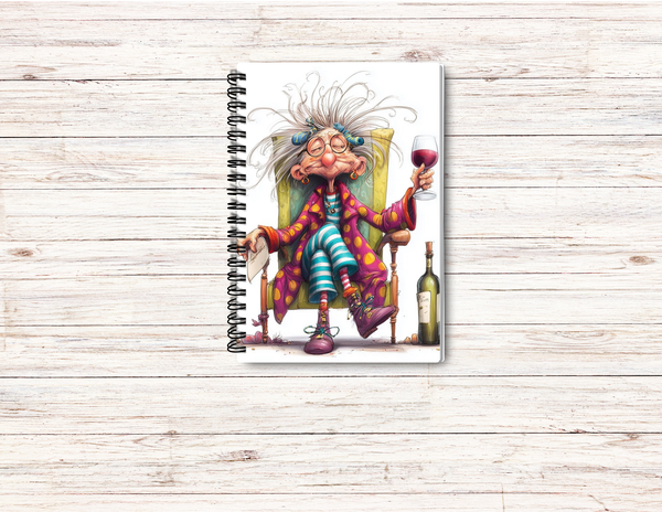 A spiral-bound notebook featuring a colorful illustration of a whimsical, wild-eyed woman in a yellow armchair holding a glass of red wine, displayed on a light wood plank background.