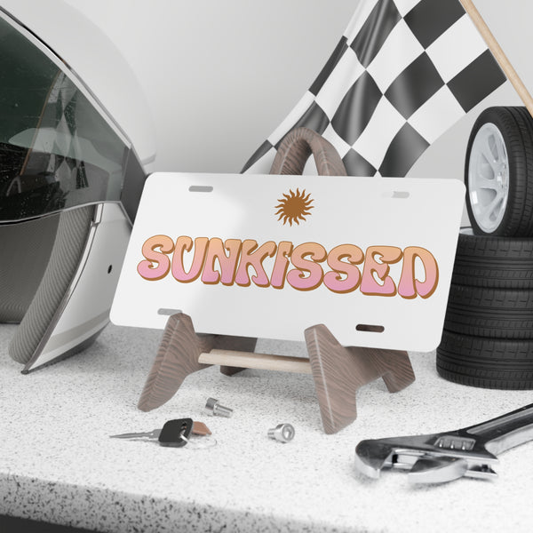 License plate with 'SUNKISSED' text on a stand, surrounded by car parts and tools.
